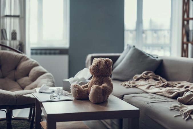 A small teddy bear sits beside a box of tissues in a softly lit living space, slightly separated from its surroundings. The scene feels quiet and emotionally heavy, photographed in a realistic editorial style with no people or distracting objects.